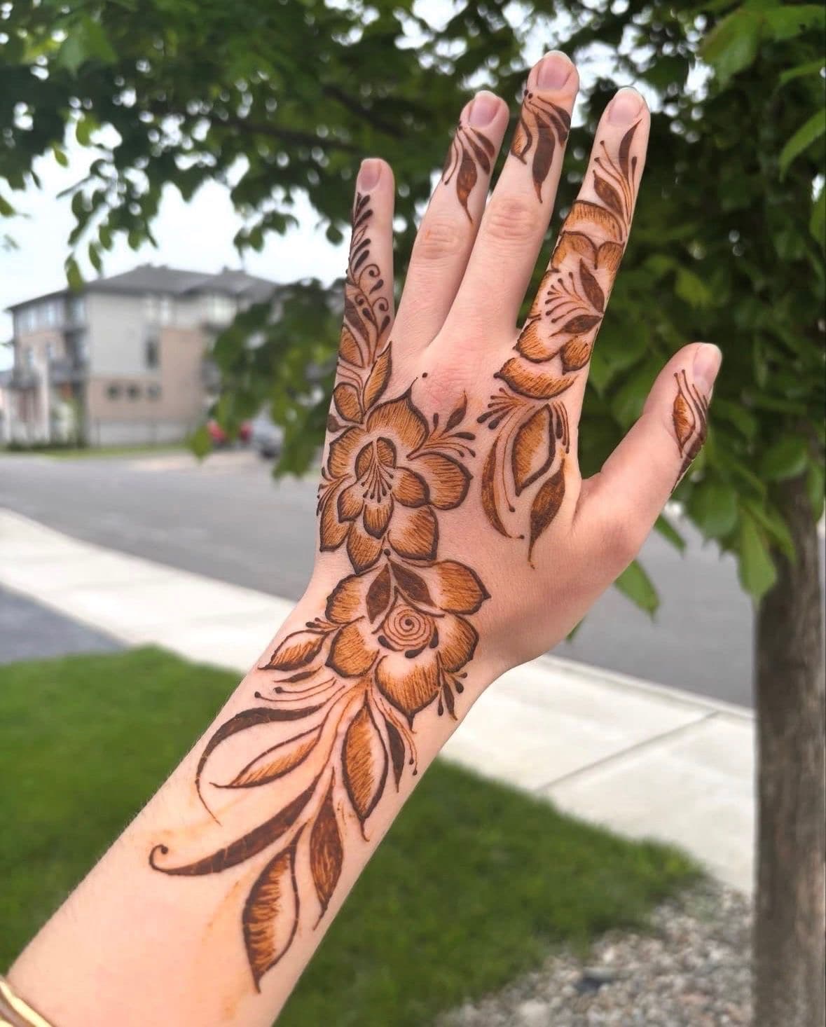 Hand raised against trees and sky, large brown henna floral on the back of the hand and wrist